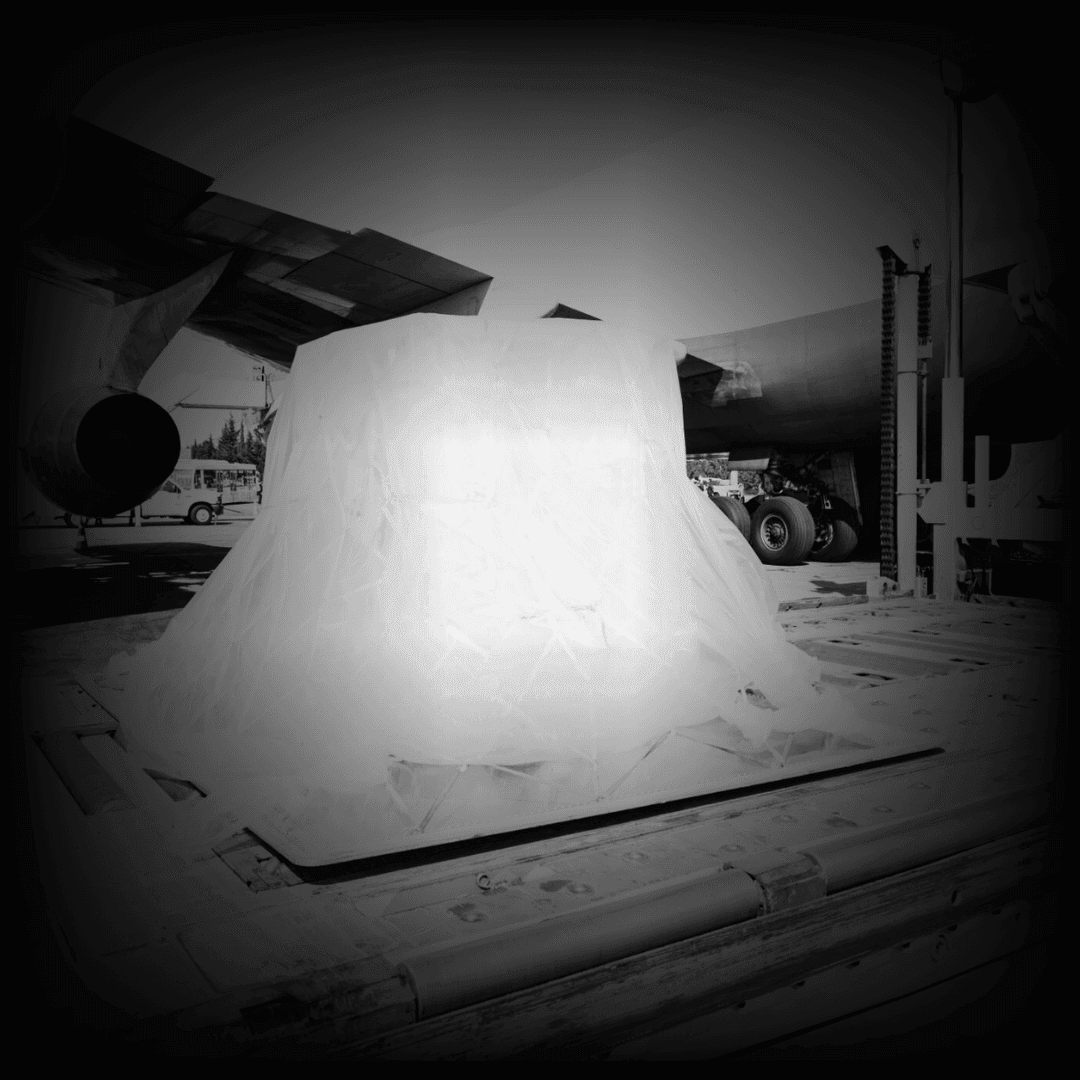 Large object wrapped in plastic sheeting next to a parked airplane on an airfield.